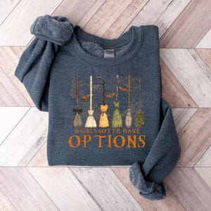 May include: A dark blue sweatshirt with the text "A Girl's Gotta Have Options" in orange. The design features several witch brooms in various colours, including brown, white, and green, with a Halloween theme.