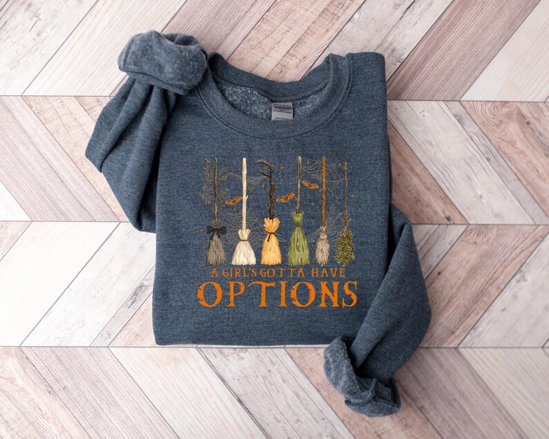 May include: A dark blue sweatshirt with the text "A Girl's Gotta Have Options" in orange. The design features several witch brooms in various colours, including brown, white, and green, with a Halloween theme.