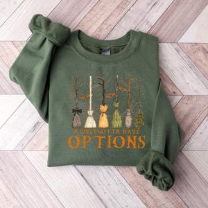 May include: An olive green sweatshirt with a design of various broomsticks with different coloured bristles. The text "A Girls Gotta Have Options" is printed in orange below the broomsticks. The sweatshirt is made of a soft material.