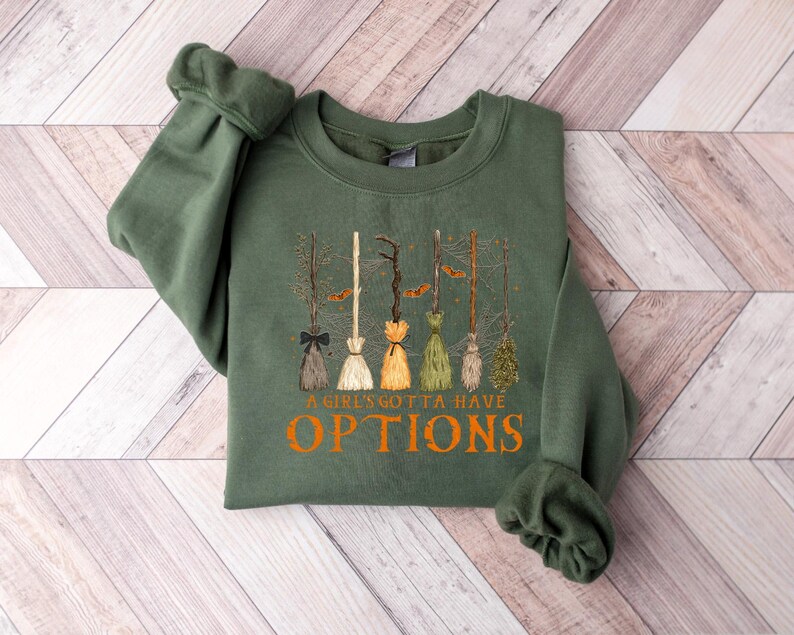 May include: An olive green sweatshirt with a design of various broomsticks with different coloured bristles. The text "A Girls Gotta Have Options" is printed in orange below the broomsticks. The sweatshirt is made of a soft material.
