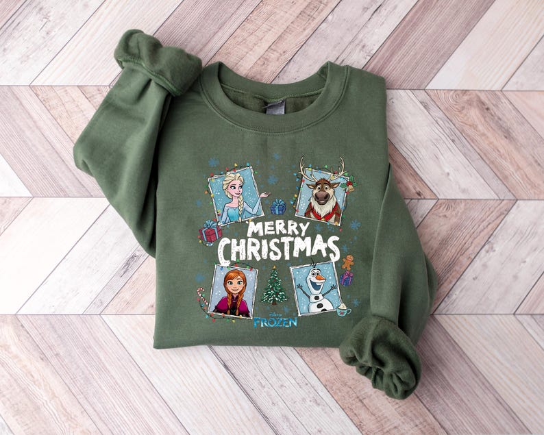Puede incluir: Una sudadera verde oliva con personajes de la pel&iacute;cula Frozen. El dise&ntilde;o incluye a Elsa, Anna, Olaf y Sven, con las palabras "Merry Christmas" y la palabra "Frozen" debajo. La sudadera est&aacute; sobre una superficie de madera.