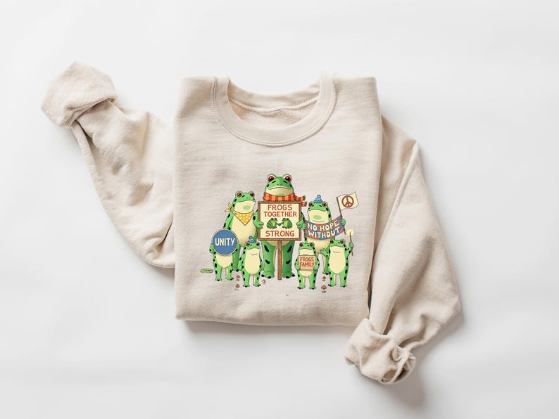 Puede incluir: Sudadera beige con un dise&ntilde;o de ranas verdes sosteniendo carteles con las frases "Unity", "Frogs Together Strong", "No Hope Without" y "Frogs Family". Las ranas llevan accesorios. Tambi&eacute;n se ve un s&iacute;mbolo de la paz.