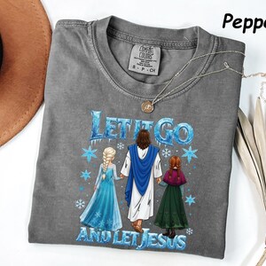 May include: A gray Comfort Colors t-shirt with the phrase "LET IT GO AND LET JESUS" in blue and white. The graphic features Jesus, Elsa, and Anna. A silver necklace is draped across the shirt.