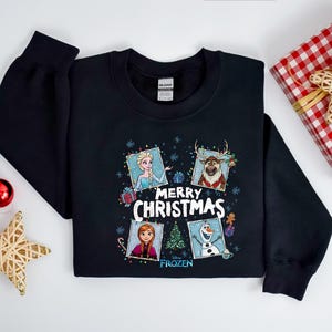Puede incluir: Sudadera negra con personajes de la pel&iacute;cula Frozen. El dise&ntilde;o incluye a Elsa, Anna, Olaf y Sven, con las palabras "Merry Christmas" y "Frozen". Tambi&eacute;n se ve un regalo envuelto y adornos.
