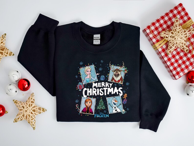Puede incluir: Sudadera negra con personajes de la pel&iacute;cula Frozen. El dise&ntilde;o incluye a Elsa, Anna, Olaf y Sven, con las palabras "Merry Christmas" y "Frozen". Tambi&eacute;n se ve un regalo envuelto y adornos.