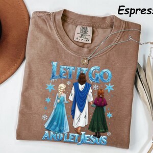 May include: Espresso-colored t-shirt with the text "LET IT GO AND LET JESUS" in blue and white. The graphic depicts Jesus with two figures in blue and green dresses. A silver necklace with a pendant is draped on the shirt.