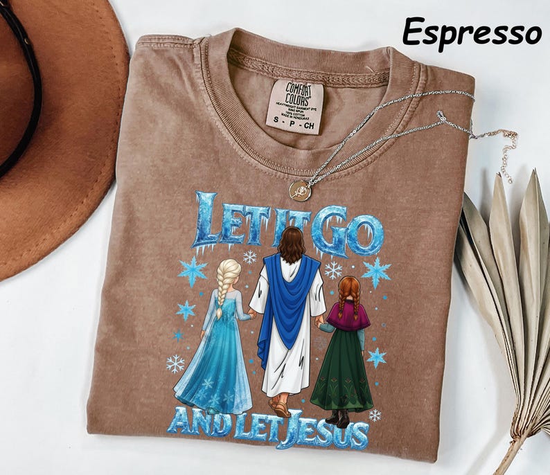 May include: Espresso-colored t-shirt with the text "LET IT GO AND LET JESUS" in blue and white. The graphic depicts Jesus with two figures in blue and green dresses. A silver necklace with a pendant is draped on the shirt.