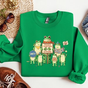 Puede incluir: Una sudadera verde brillante con un dise&ntilde;o de ranas. El dise&ntilde;o muestra varias ranas de dibujos animados sosteniendo carteles con lemas como "Frogs Together Strong" y "No Hope Without". Algunas ranas llevan accesorios y se incluye un s&iacute;mbolo de la paz.
