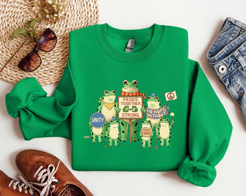 Puede incluir: Una sudadera verde brillante con un dise&ntilde;o de ranas. El dise&ntilde;o muestra varias ranas de dibujos animados sosteniendo carteles con lemas como "Frogs Together Strong" y "No Hope Without". Algunas ranas llevan accesorios y se incluye un s&iacute;mbolo de la paz.