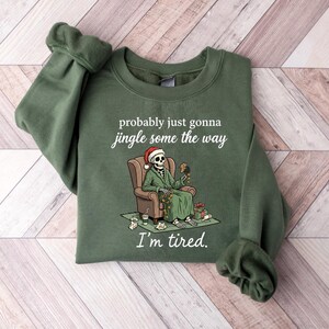 K&ouml;nnte beinhalten: Gr&uuml;nes Sweatshirt mit dem Text "probably just gonna jingle some the way" und "I'm tired." Die Grafik zeigt ein Skelett mit Weihnachtsm&uuml;tze und Anzug, das in einem Stuhl sitzt. Geschenke und eine Kaffeetasse liegen auf dem Boden.