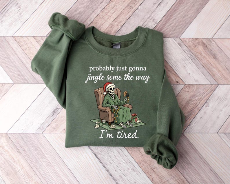 K&ouml;nnte beinhalten: Gr&uuml;nes Sweatshirt mit dem Text "probably just gonna jingle some the way" und "I'm tired." Die Grafik zeigt ein Skelett mit Weihnachtsm&uuml;tze und Anzug, das in einem Stuhl sitzt. Geschenke und eine Kaffeetasse liegen auf dem Boden.