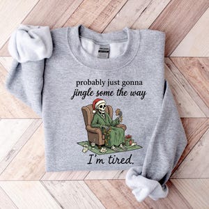 K&ouml;nnte beinhalten: Hellgraues Sweatshirt mit dem Text "probably just gonna jingle some the way" und "I'm tired." Ein Skelett mit Weihnachtsm&uuml;tze und gr&uuml;nem Mantel sitzt in einem braunen Sessel und h&auml;lt Dekorationen. Weihnachts-Themenbekleidung.