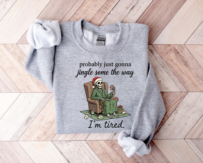K&ouml;nnte beinhalten: Hellgraues Sweatshirt mit dem Text "probably just gonna jingle some the way" und "I'm tired." Ein Skelett mit Weihnachtsm&uuml;tze und gr&uuml;nem Mantel sitzt in einem braunen Sessel und h&auml;lt Dekorationen. Weihnachts-Themenbekleidung.