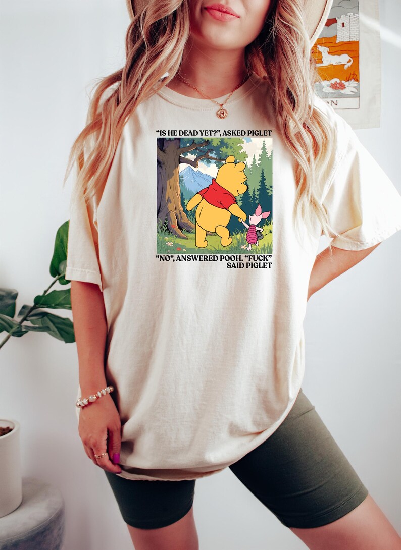 Puede incluir: Camiseta color crema con una ilustraci&oacute;n de dibujos animados de Winnie the Pooh y Piglet. La imagen incluye el texto "IS HE DEAD YET?" y "NO", ANSWERED POOH. "FUCK" SAID PIGLET". La camiseta es una prenda casual.