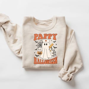 May include: Beige sweatshirt with the text "Pappy Halloween" in orange, featuring a cartoon ghost dressed as a nurse holding a pumpkin and a syringe. The design includes bats and spiderwebs, perfect for Halloween.