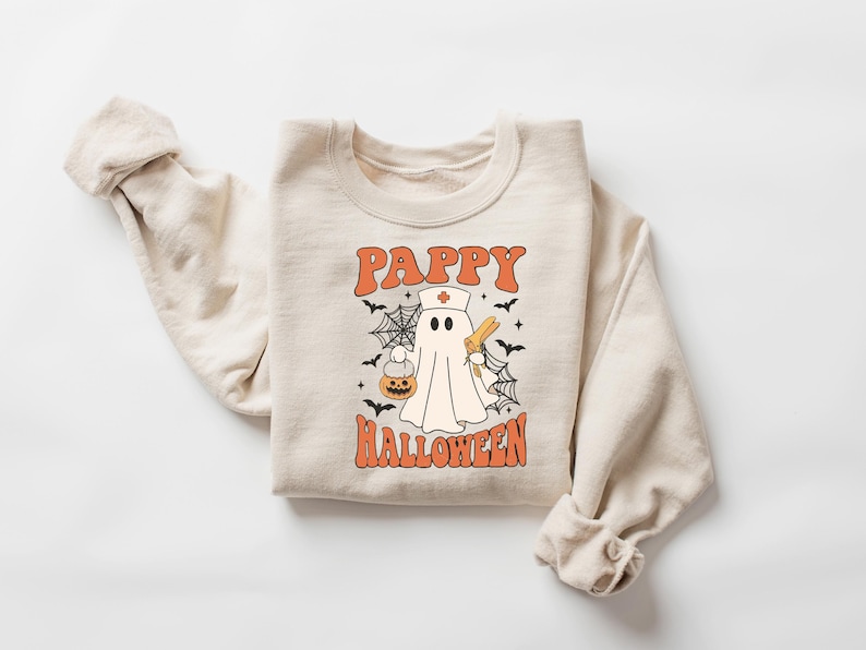 May include: Beige sweatshirt with the text "Pappy Halloween" in orange, featuring a cartoon ghost dressed as a nurse holding a pumpkin and a syringe. The design includes bats and spiderwebs, perfect for Halloween.