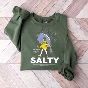 May include: An olive green sweatshirt with a graphic of a person under a blue umbrella, with the word "SALTY" in white. The person is wearing a yellow dress and is sprinkling salt. The text "All those flavors and you choose to be" is also present.