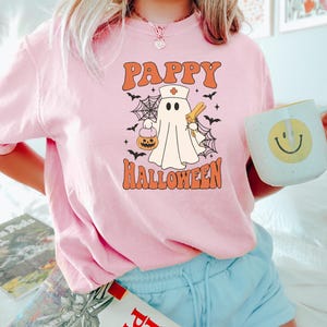 May include: Pink t-shirt with the text "Pappy Halloween" in orange. The graphic features a ghost wearing a nurse's hat, holding a syringe and a pumpkin. Black bats and spiderwebs surround the ghost. A white mug with a yellow smiley face is held in the hand.