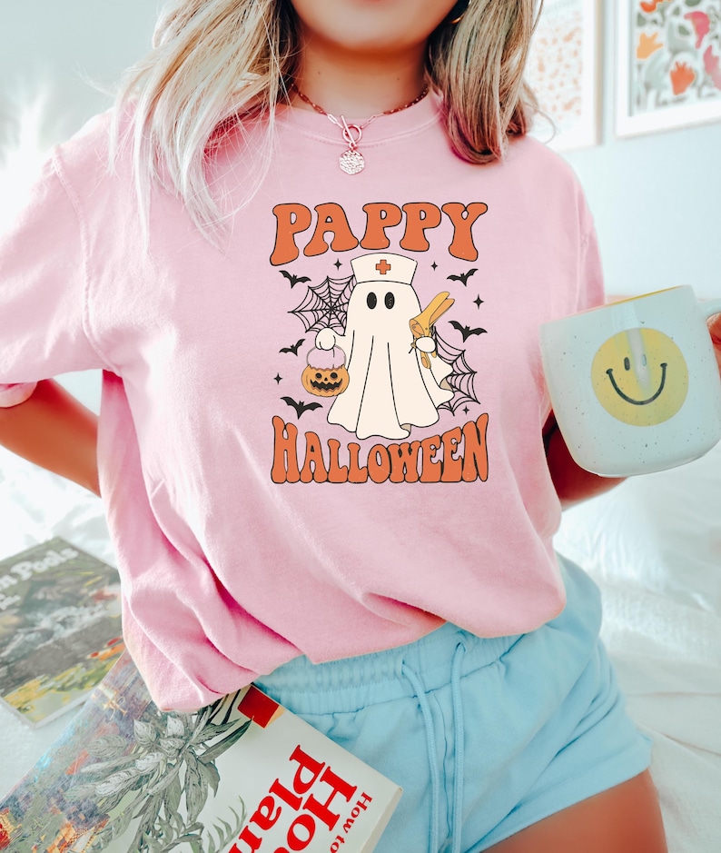 May include: Pink t-shirt with the text "Pappy Halloween" in orange. The graphic features a ghost wearing a nurse's hat, holding a syringe and a pumpkin. Black bats and spiderwebs surround the ghost. A white mug with a yellow smiley face is held in the hand.