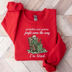 K&ouml;nnte beinhalten: Rotes Sweatshirt mit humorvoller Grafik. Das Design zeigt ein Skelett mit Weihnachtsm&uuml;tze und gr&uuml;nem Mantel, das in einem Stuhl sitzt. Der Text lautet "probably just gonna jingle some the way, I'm tired."