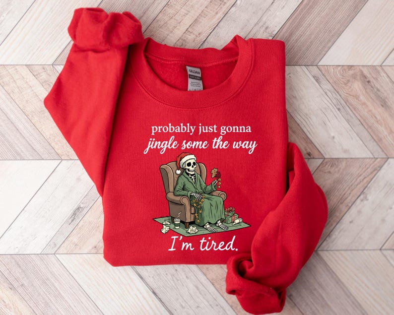 K&ouml;nnte beinhalten: Rotes Sweatshirt mit humorvoller Grafik. Das Design zeigt ein Skelett mit Weihnachtsm&uuml;tze und gr&uuml;nem Mantel, das in einem Stuhl sitzt. Der Text lautet "probably just gonna jingle some the way, I'm tired."