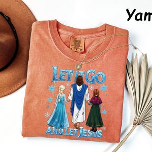 May include: A coral-colored t-shirt with the phrase "Let It Go And Let Jesus" in blue and white. The graphic depicts Jesus holding hands with two animated characters. A gold necklace and a brown hat are also visible.