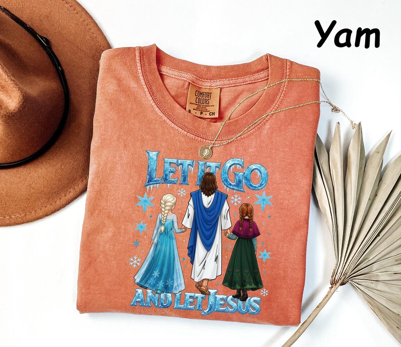May include: A coral-colored t-shirt with the phrase "Let It Go And Let Jesus" in blue and white. The graphic depicts Jesus holding hands with two animated characters. A gold necklace and a brown hat are also visible.
