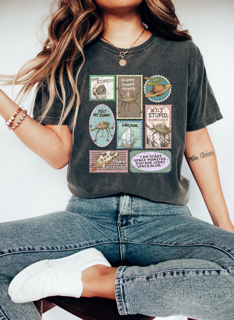 May include: Dark gray t-shirt featuring a collection of whimsical postage stamp designs. The stamps display various space-themed illustrations and phrases like "Why Stupid?" and "I am scary space monster."