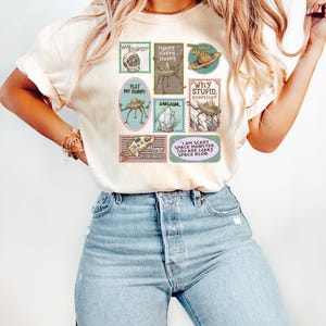 May include: Cream-colored t-shirt with a collection of whimsical, postage stamp-style graphics. The stamps feature various designs, including a space helmet, a robot, and the text "Why Stupid?" and "I am scary space monster."