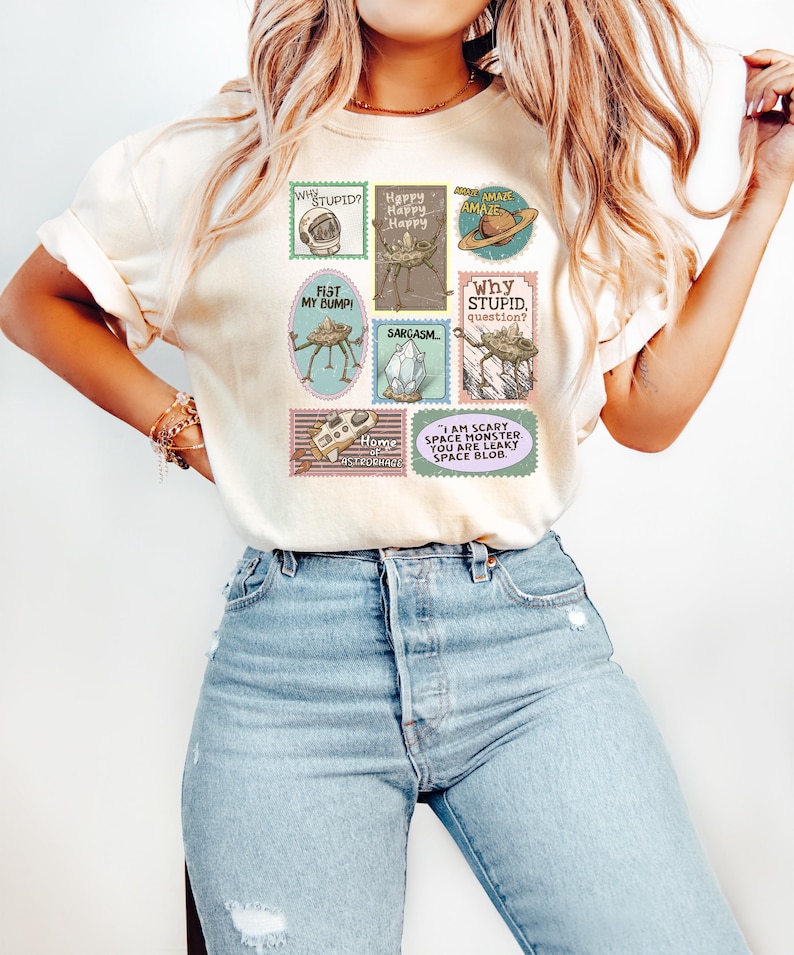 May include: Cream-colored t-shirt with a collection of whimsical, postage stamp-style graphics. The stamps feature various designs, including a space helmet, a robot, and the text "Why Stupid?" and "I am scary space monster."