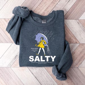 May include: Dark gray sweatshirt with a graphic of a person under a purple umbrella, wearing a yellow dress, and pouring salt. The word "SALTY" is printed in large white letters below the graphic. Text above the graphic reads "All the flavors and you choose to be."