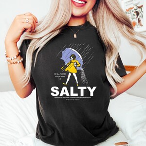 May include: Black t-shirt with a graphic of a person under a blue umbrella, with salt pouring down. The word "SALTY" is printed in large white letters below the graphic. Text on the shirt reads "All these flavors and you choose to be."