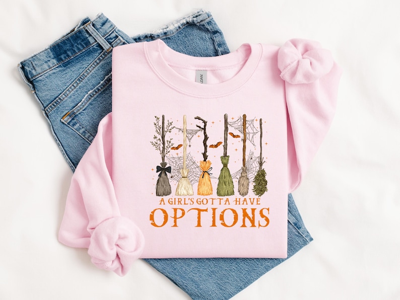 May include: A pale pink sweatshirt with the slogan "A Girl's Gotta Have Options" in orange. The design shows various witch brooms, spiderwebs, and bats, ideal for Halloween. The sweatshirt is placed on top of blue denim jeans.