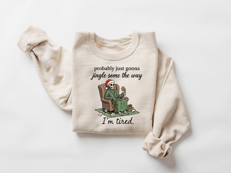 K&ouml;nnte beinhalten: Beiges Sweatshirt mit dem Text "probably just gonna jingle some the way, I'm tired." Die Grafik zeigt ein Skelett mit Weihnachtsm&uuml;tze, das in einem Stuhl sitzt, umgeben von Geschenken. Ein humorvolles Design zum Thema Weihnachten.