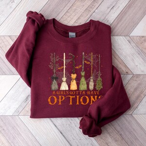 May include: A burgundy sweatshirt featuring a graphic of several witch brooms in different colours. The text "A GIRL'S GOTTA HAVE OPTIONS" is printed in orange below the brooms. The sweatshirt is folded on a light wood-grain surface.