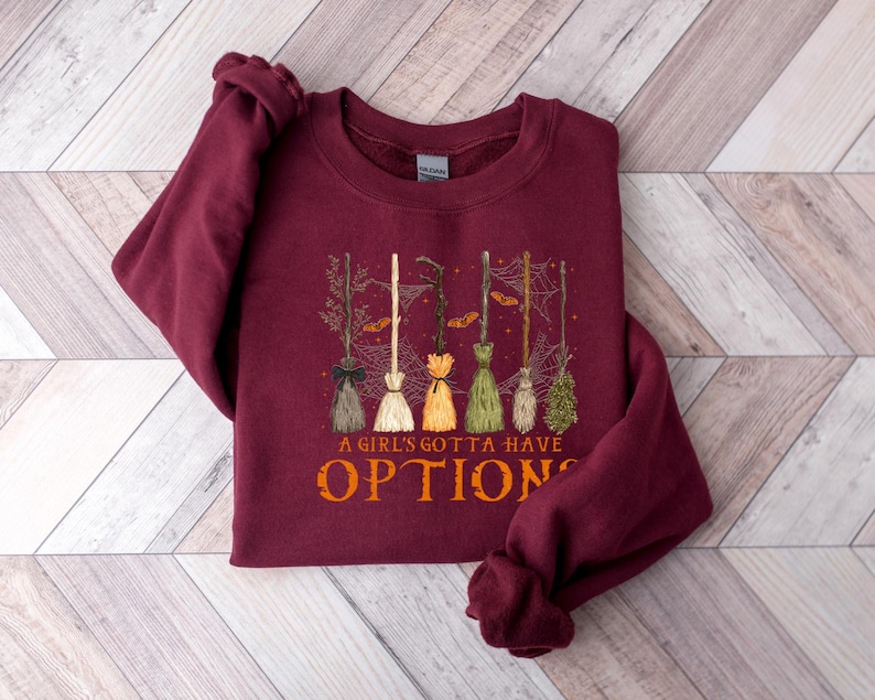 May include: A burgundy sweatshirt featuring a graphic of several witch brooms in different colours. The text "A GIRL'S GOTTA HAVE OPTIONS" is printed in orange below the brooms. The sweatshirt is folded on a light wood-grain surface.