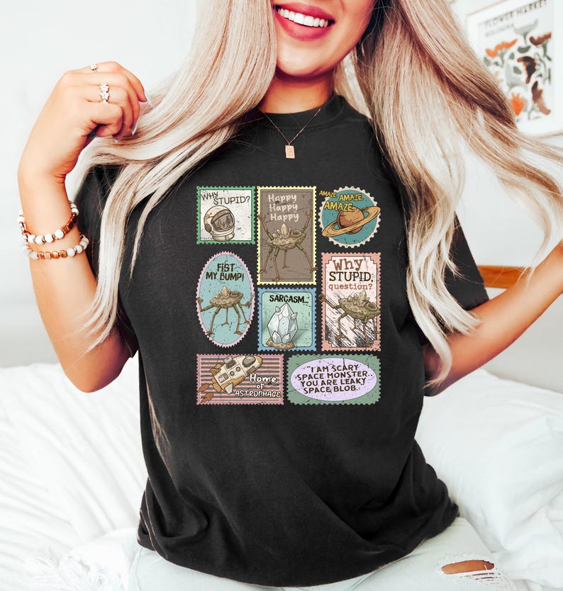 May include: Black t-shirt featuring a collection of whimsical postage stamp designs. The stamps include phrases like "Why Stupid?" and "I am Scary Space Monster." The shirt has a crew neck and short sleeves.