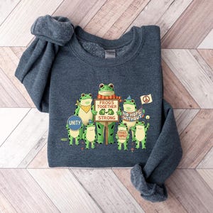 Puede incluir: Sudadera azul oscuro con un gr&aacute;fico de ranas de dibujos animados verdes que sostienen carteles con mensajes como "Frogs Together Strong" y "No Hope Without". Tambi&eacute;n se muestra la palabra "Unity". La sudadera tiene cuello redondo.