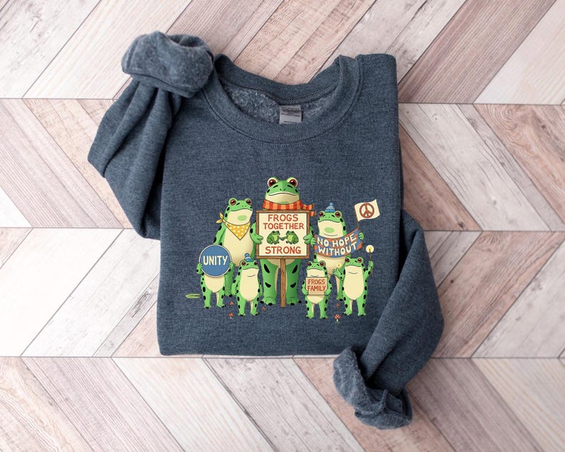 Puede incluir: Sudadera azul oscuro con un gr&aacute;fico de ranas de dibujos animados verdes que sostienen carteles con mensajes como "Frogs Together Strong" y "No Hope Without". Tambi&eacute;n se muestra la palabra "Unity". La sudadera tiene cuello redondo.