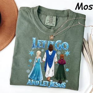 May include: Moss green t-shirt with the text "Let It Go and Let Jesus" in blue. The graphic features three figures in a winter scene. A gold necklace is draped across the shirt.
