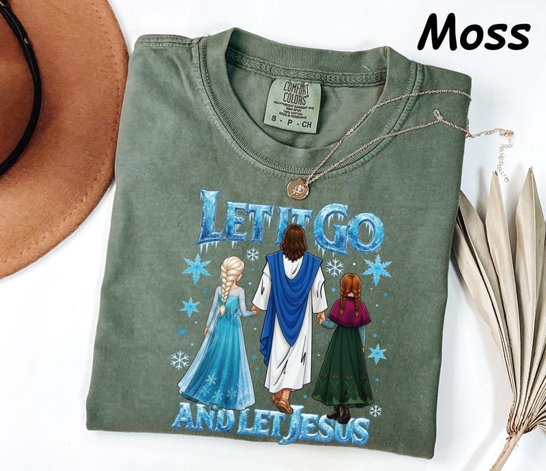 May include: Moss green t-shirt with the text "Let It Go and Let Jesus" in blue. The graphic features three figures in a winter scene. A gold necklace is draped across the shirt.