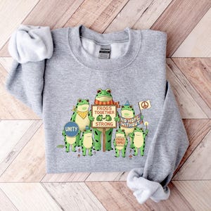 Puede incluir: Una sudadera gris jaspeada con un gr&aacute;fico de ranas verdes sosteniendo carteles. Los carteles dicen "Frogs Together Strong", "No Hope Without" y "Frogs Family". Una rana sostiene un cartel que dice "Unity". La sudadera tiene cuello redondo y mangas largas.