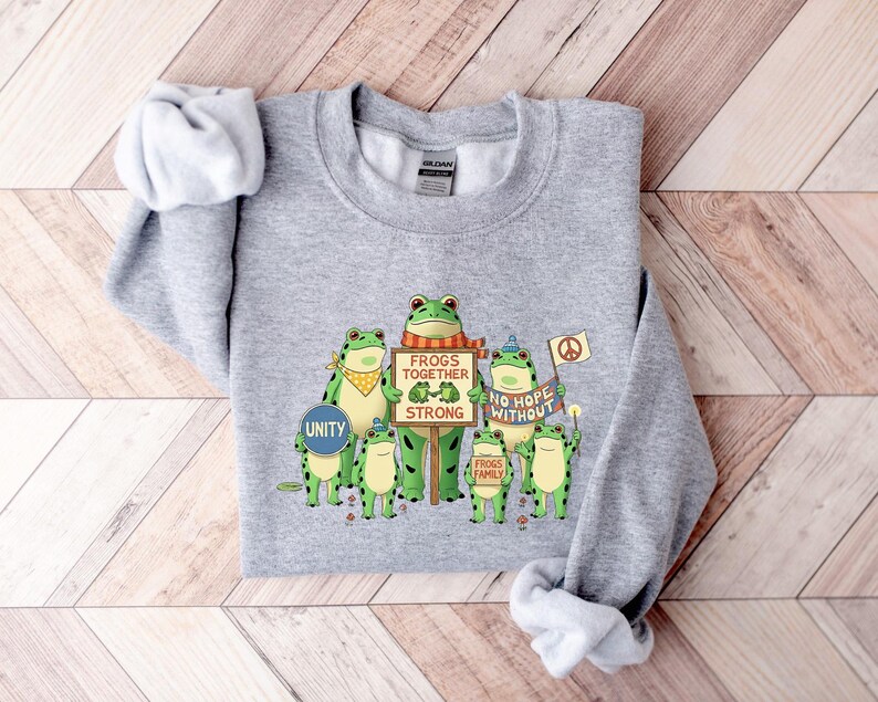 Puede incluir: Una sudadera gris jaspeada con un gr&aacute;fico de ranas verdes sosteniendo carteles. Los carteles dicen "Frogs Together Strong", "No Hope Without" y "Frogs Family". Una rana sostiene un cartel que dice "Unity". La sudadera tiene cuello redondo y mangas largas.