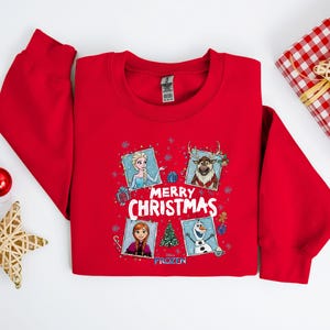 Puede incluir: Una sudadera roja con un dise&ntilde;o festivo con personajes de la pel&iacute;cula Frozen. El dise&ntilde;o incluye a Elsa, Anna, Olaf y Sven, con las palabras "Merry Christmas" y un &aacute;rbol de Navidad. Tambi&eacute;n se ven un regalo envuelto y adornos.