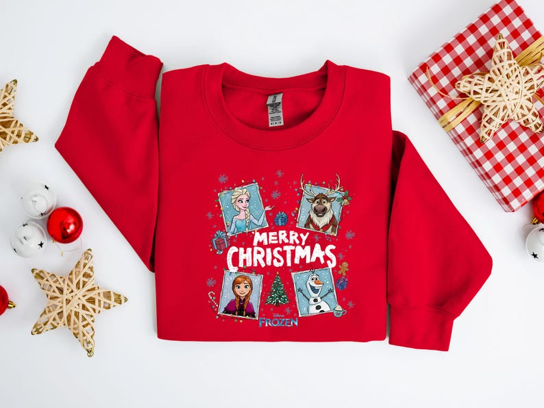Puede incluir: Una sudadera roja con un dise&ntilde;o festivo con personajes de la pel&iacute;cula Frozen. El dise&ntilde;o incluye a Elsa, Anna, Olaf y Sven, con las palabras "Merry Christmas" y un &aacute;rbol de Navidad. Tambi&eacute;n se ven un regalo envuelto y adornos.
