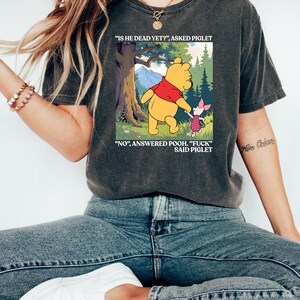 Puede incluir: Camiseta gris oscuro con una imagen de dibujos animados de Winnie the Pooh y Piglet. La imagen muestra a Pooh y Piglet caminando en un bosque con el texto "IS HE DEAD YET?" y "NO, ANSWERED POOH. 'FUCK' SAID PIGLET."