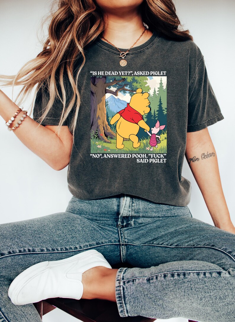 Puede incluir: Camiseta gris oscuro con una imagen de dibujos animados de Winnie the Pooh y Piglet. La imagen muestra a Pooh y Piglet caminando en un bosque con el texto "IS HE DEAD YET?" y "NO, ANSWERED POOH. 'FUCK' SAID PIGLET."