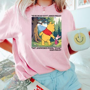 Puede incluir: Sudadera rosa claro con un gr&aacute;fico de Winnie the Pooh y Piglet con texto. La imagen muestra a Pooh y Piglet en un entorno forestal con el texto "IS HE DEAD YET?" y "NO, ANSWERED POOH. 'FUCK' SAID PIGLET."