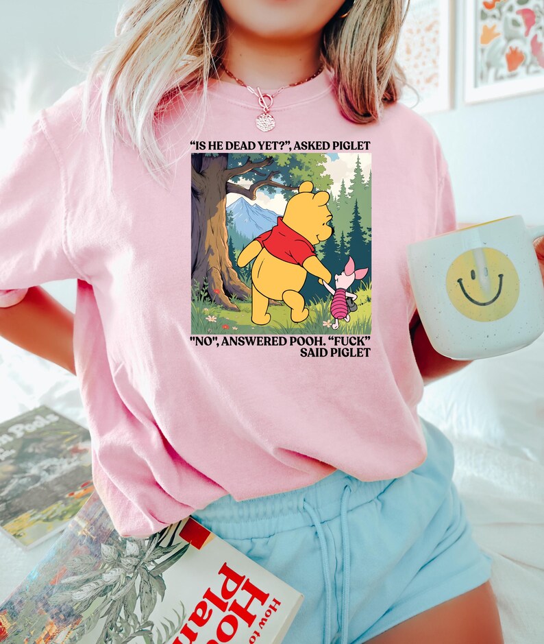 Puede incluir: Sudadera rosa claro con un gr&aacute;fico de Winnie the Pooh y Piglet con texto. La imagen muestra a Pooh y Piglet en un entorno forestal con el texto "IS HE DEAD YET?" y "NO, ANSWERED POOH. 'FUCK' SAID PIGLET."
