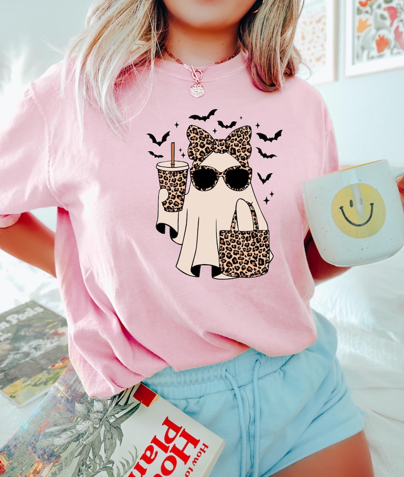 May include: Pink t-shirt featuring a cartoon ghost wearing sunglasses, a bow, and carrying a leopard print bag and drink. Black bats are flying above the ghost. A white mug with a yellow smiley face is held in the hand.